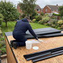 Roofer rolling water-based deck adhesive onto an OSB garden room roof deck with the one-piece EPDM membrane folded back and black metal edge trims ready - cold applied, no heat