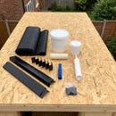 Garden room rubber roof kit contents laid out on an OSB deck: EPDM membrane, water-based deck adhesive, roller, edge mastic, black metal edge trims, corners and screws