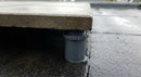 AI-generated illustrative scene (not a photograph of the actual product): close three-quarter view of a paving slab corner resting on a pedestal support, showing the raised gap with rainwater draining beneath onto the roof membrane.