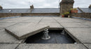 AI-generated illustrative scene (not a photograph of the actual product): an overcast UK roof terrace with square paving slabs raised on adjustable pedestal supports above a dark flat roof membrane, one slab lifted at the edge to show a support and the drainage gap beneath.