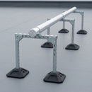 Row of galvanised H-frame rooftop supports on rubber feet carrying an insulated pipe run across a flat roof